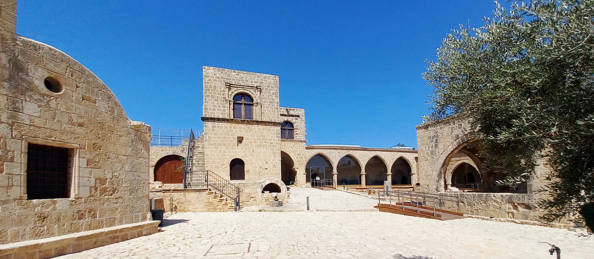 The paved courtyard of Ayia Napa Monastery under a clear blue sky, with the two-storey Venetian building and its arched upper window in the centre, a row of stone arches running off to the right and an olive tree framing the right edge