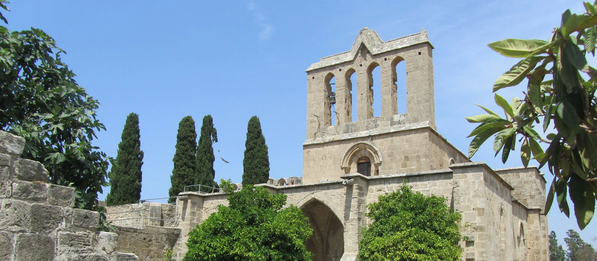 The Gothic church of Bellapais Abbey with its open stone belfry, seen from the forecourt between orange trees, with four tall cypresses against a clear blue sky