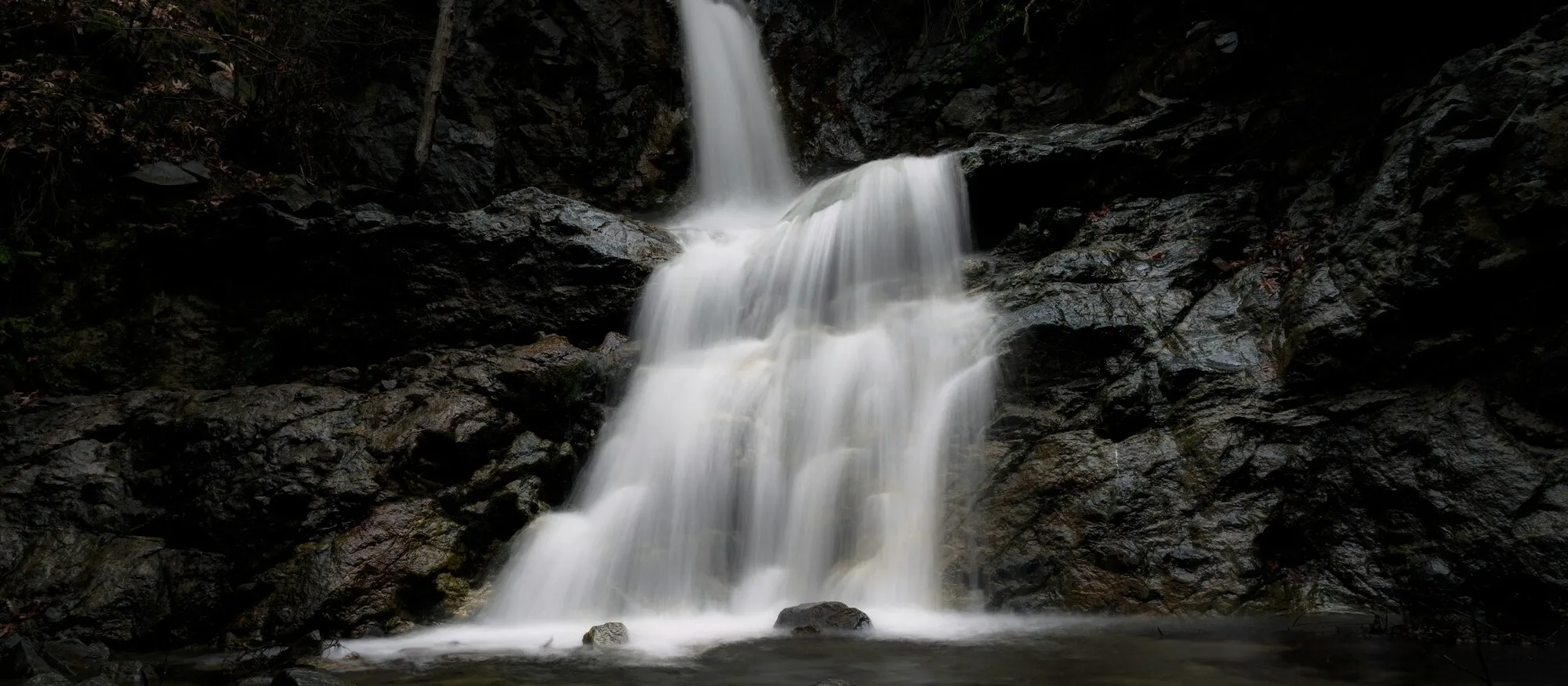 Long-exposure photograph of a stepped forest waterfall falling over dark wet rock into a shallow pool, illustrative of a Troodos mountain fall
