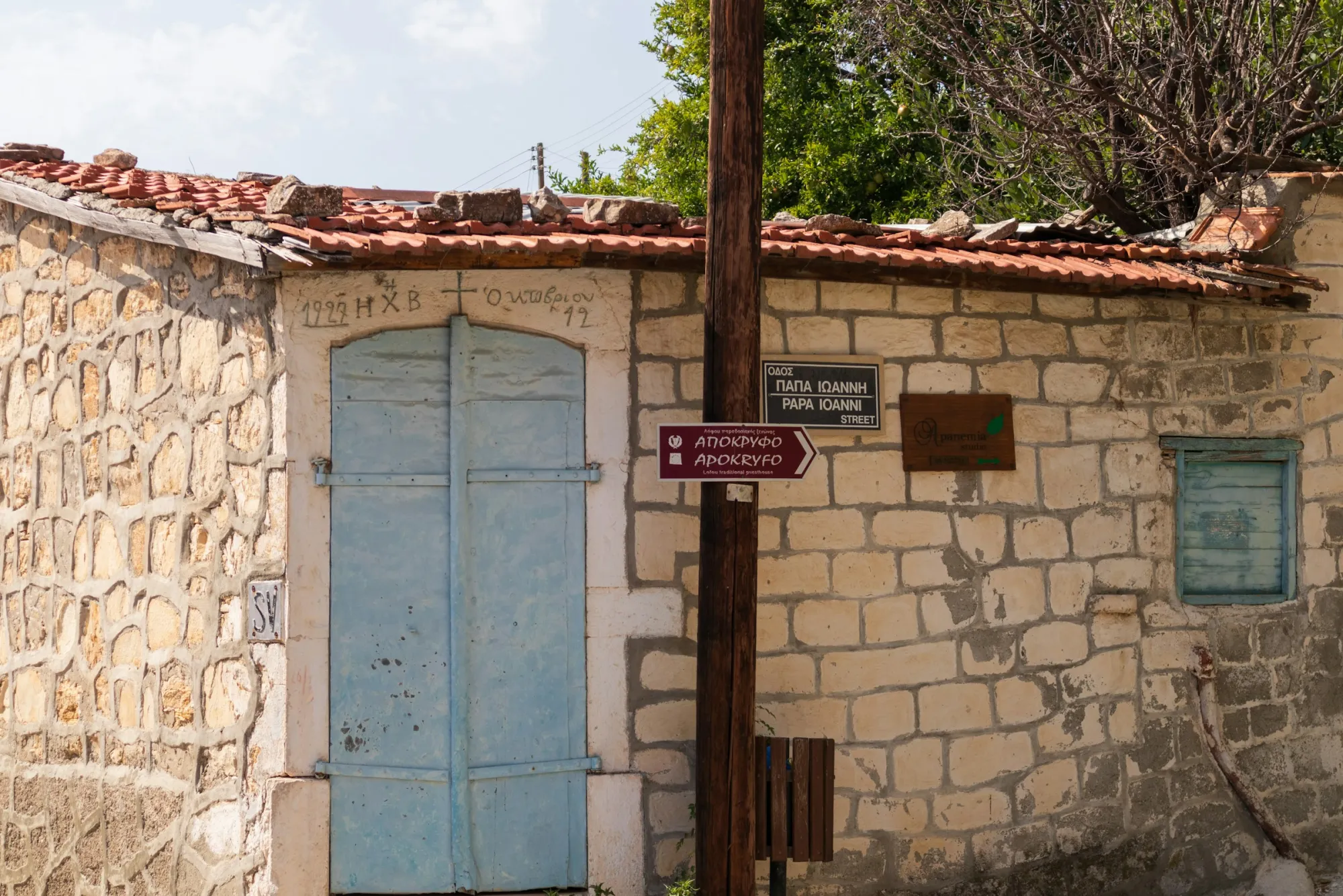 A bilingual street sign on a stone village house in Cyprus, reading ΟΔΟΣ ΠΑΠΑ ΙΩΑΝΝΗ in Greek above PAPA IOANNI STREET in Latin script