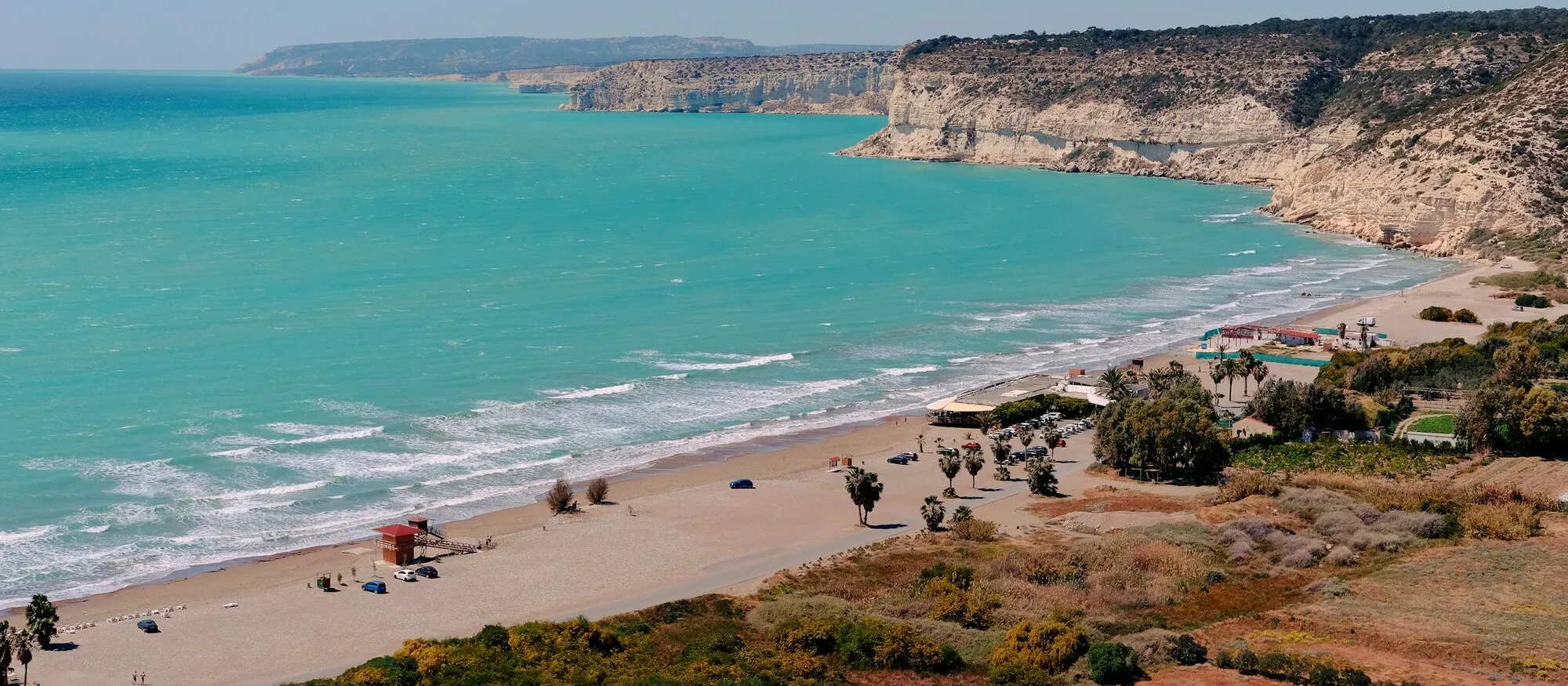 A long sandy beach curving below pale limestone cliffs on the south coast of Cyprus, with turquoise sea and a lifeguard hut on the sand