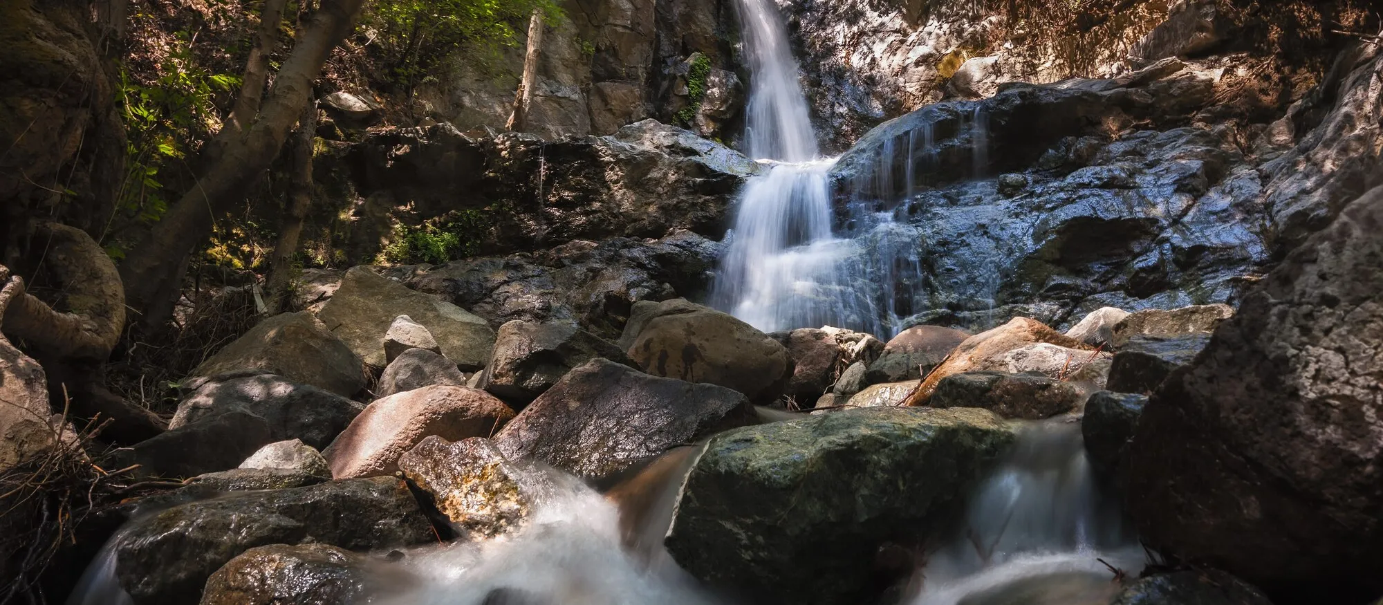 Long-exposure photograph of the Mesa Potamos waterfall in the Troodos forest, water falling over dark rock into a boulder-filled stream