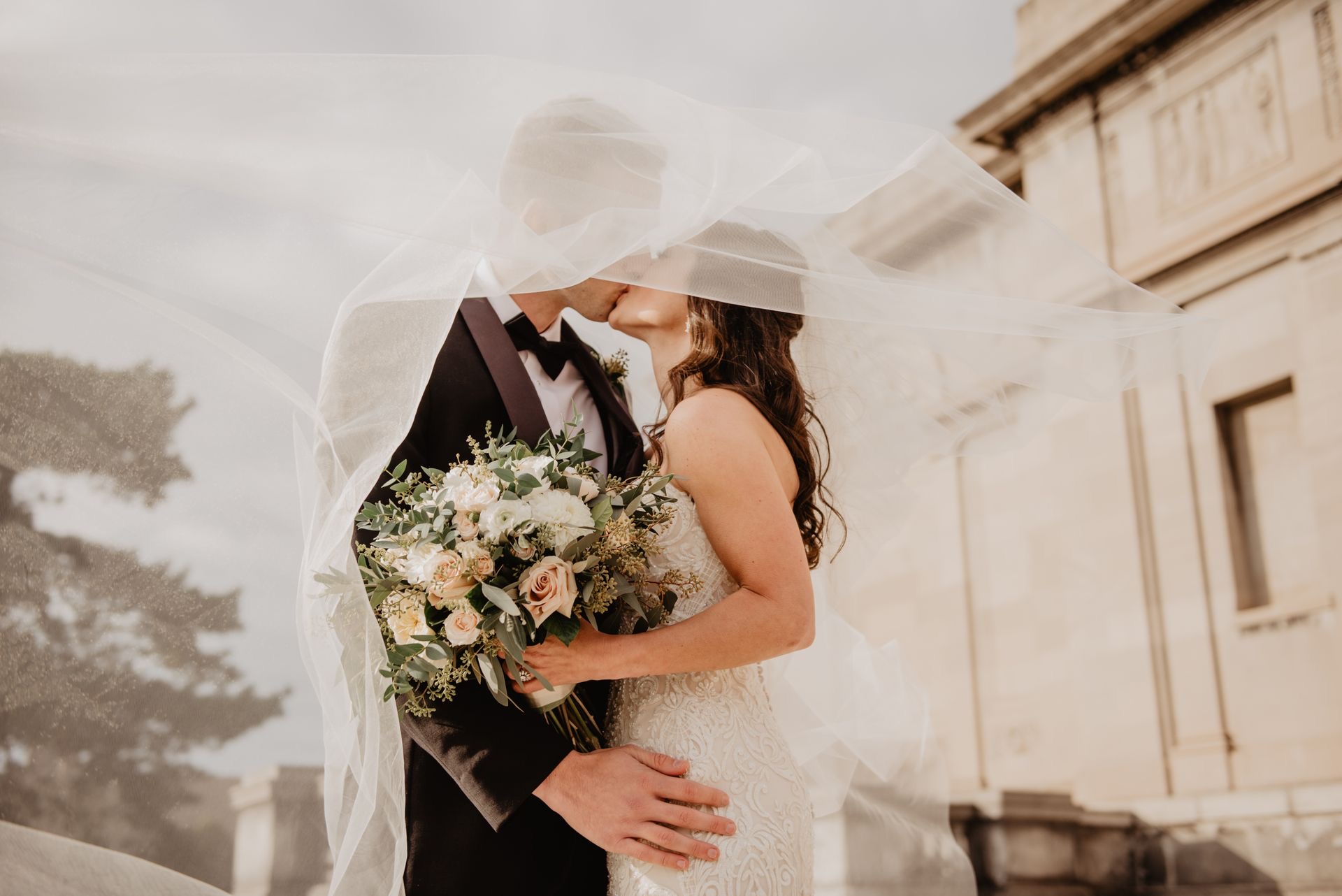 Bride and groom kissing beneath a windblown veil outside a stone building