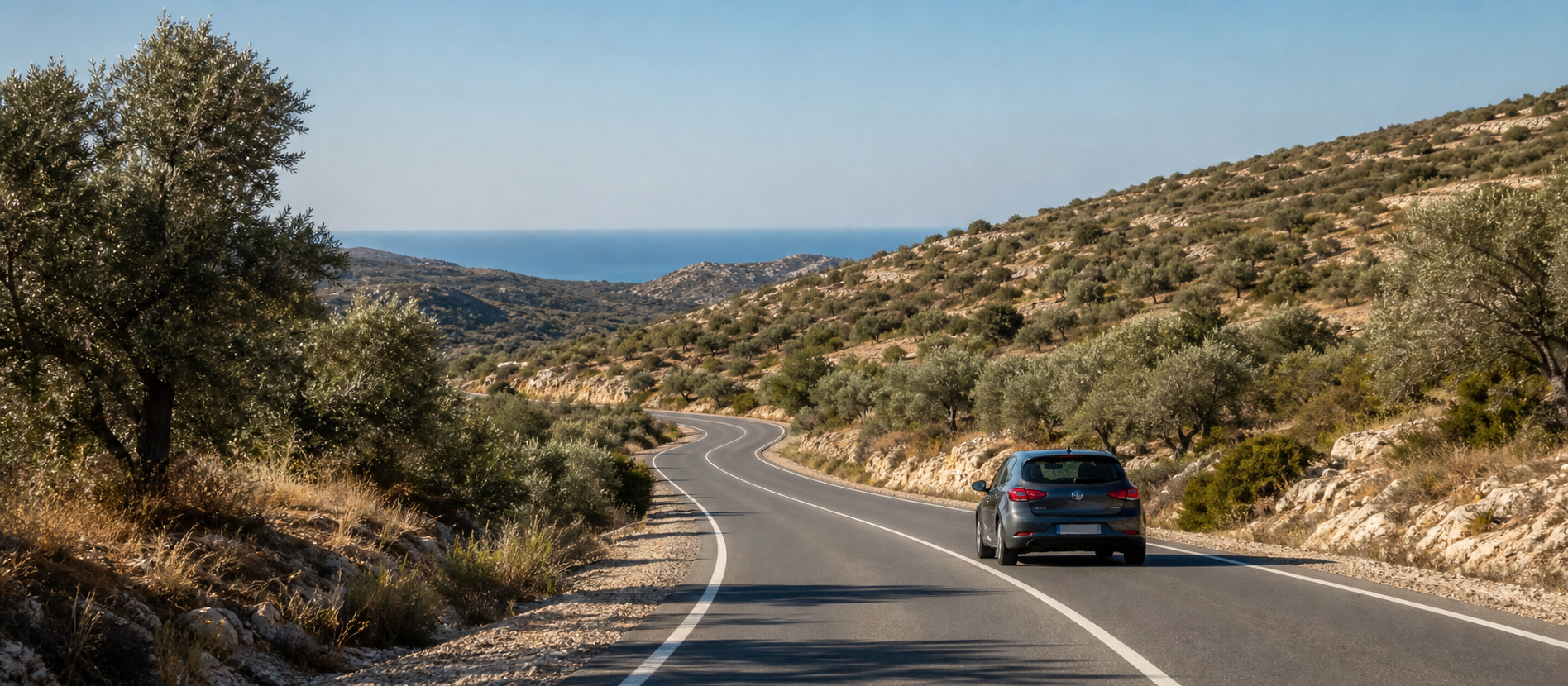 A car travelling on a quiet winding road through a Mediterranean hillside towards the sea