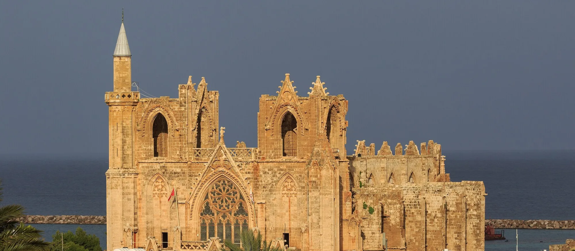 The Gothic west front of Lala Mustafa Pasha Mosque in Famagusta, once St Nicholas Cathedral, glowing honey-coloured in late sun with a single minaret on the left, the harbour breakwater and a dark sea behind and palm trees in the foreground