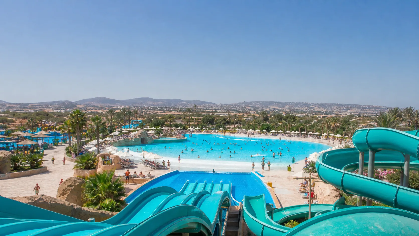 Turquoise water slides overlooking a large blue wave pool in a sunny Mediterranean water park