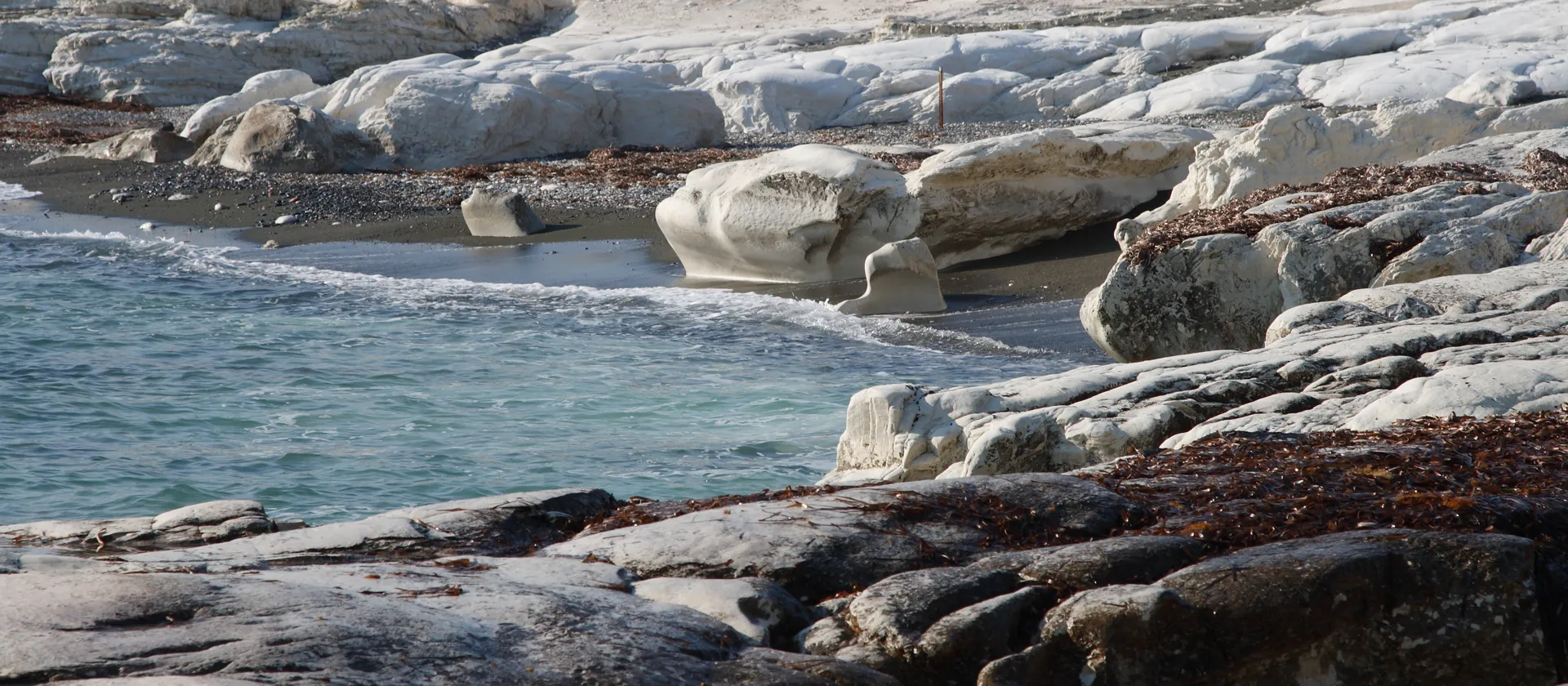 A small cove at Governor's Beach in Cyprus, with turquoise water washing onto dark sand between sculpted white chalk rocks and flat grey rock shelves in the foreground