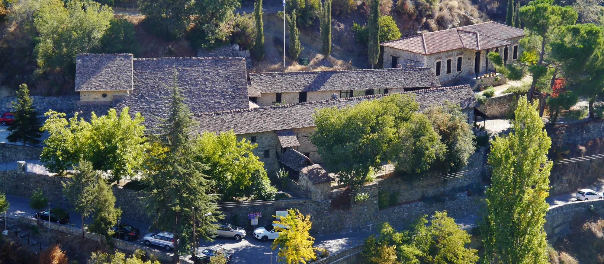 The stone-roofed Monastery of Agios Ioannis Lampadistis at Kalopanagiotis seen from above, its long grey roofs among cypresses and autumn trees, with a small car park and road beside it