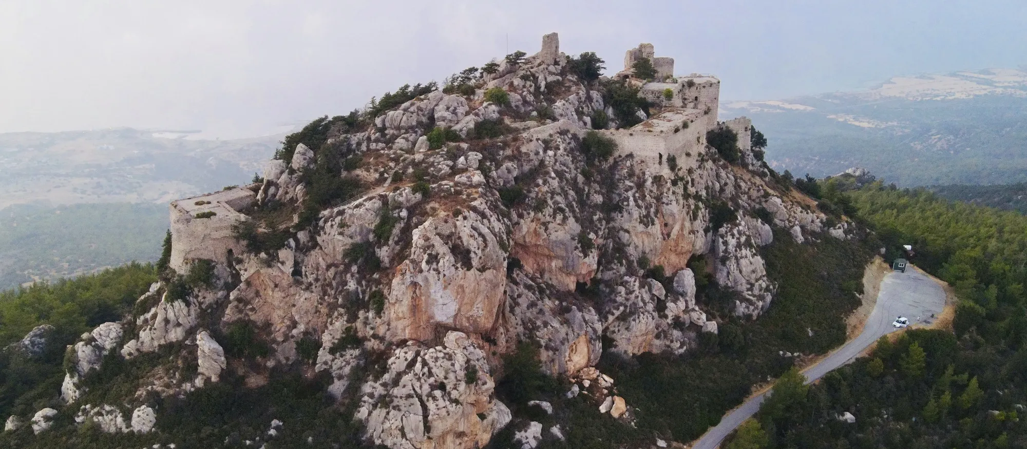 Aerial view of Kantara Castle's ruined walls and towers along the top of a pale limestone crag above pine forest, with the narrow access road curving up to a small car park on the right and the plain and sea fading into haze behind