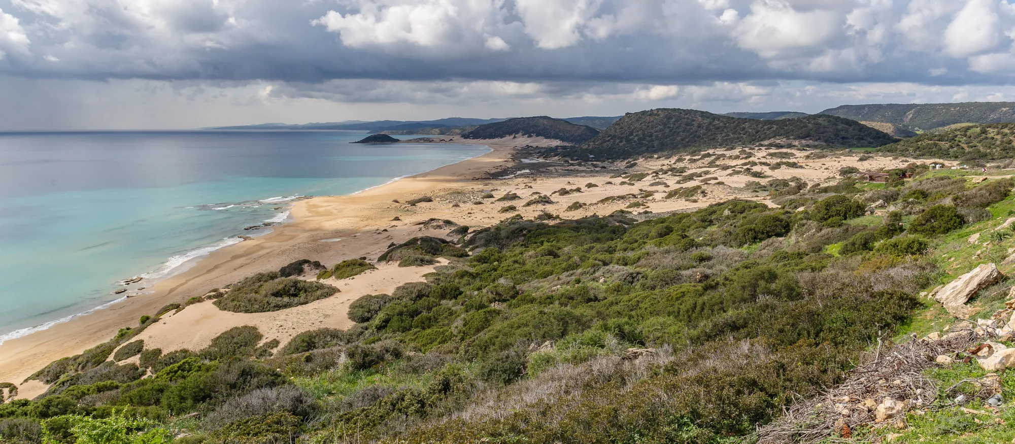 Golden Beach on the Karpas peninsula seen from a scrub-covered hillside, a long curve of pale sand and dunes running beside turquoise water towards low green hills under heavy white clouds