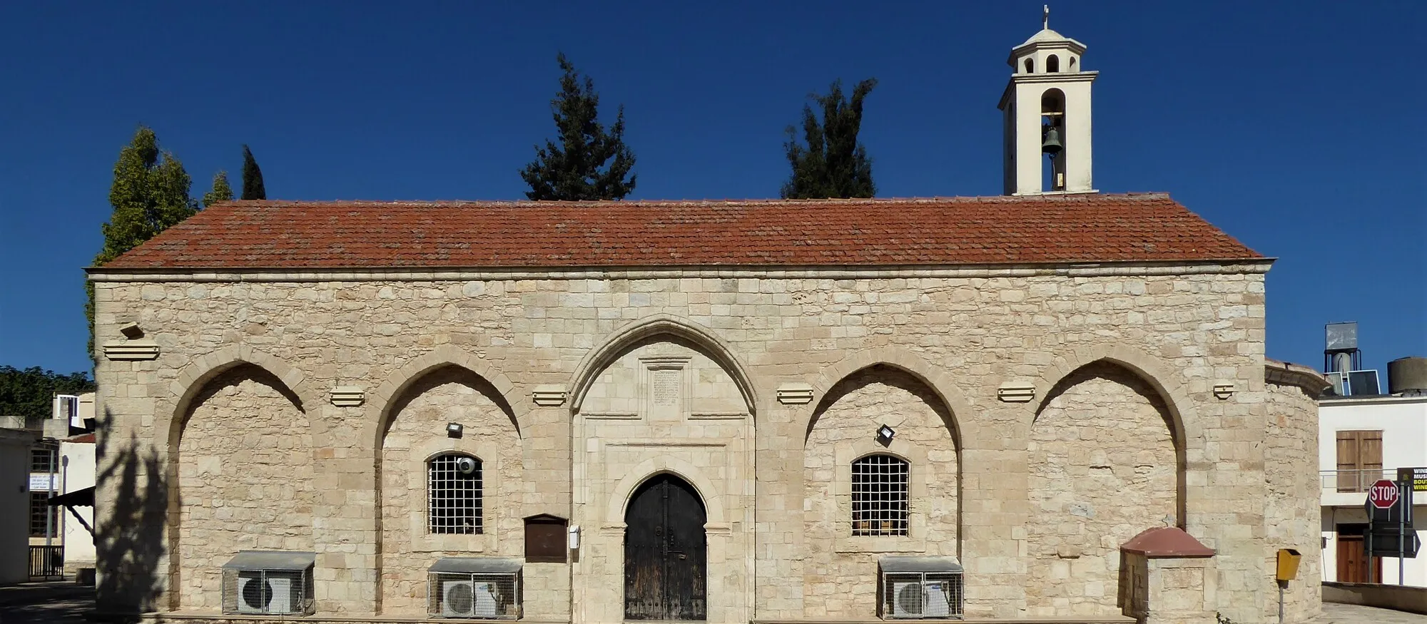 The long stone facade of Panagia Evangelistria church in Kathikas, with arched bays, a red tiled roof, a white bell tower and cypress trees against a deep blue sky
