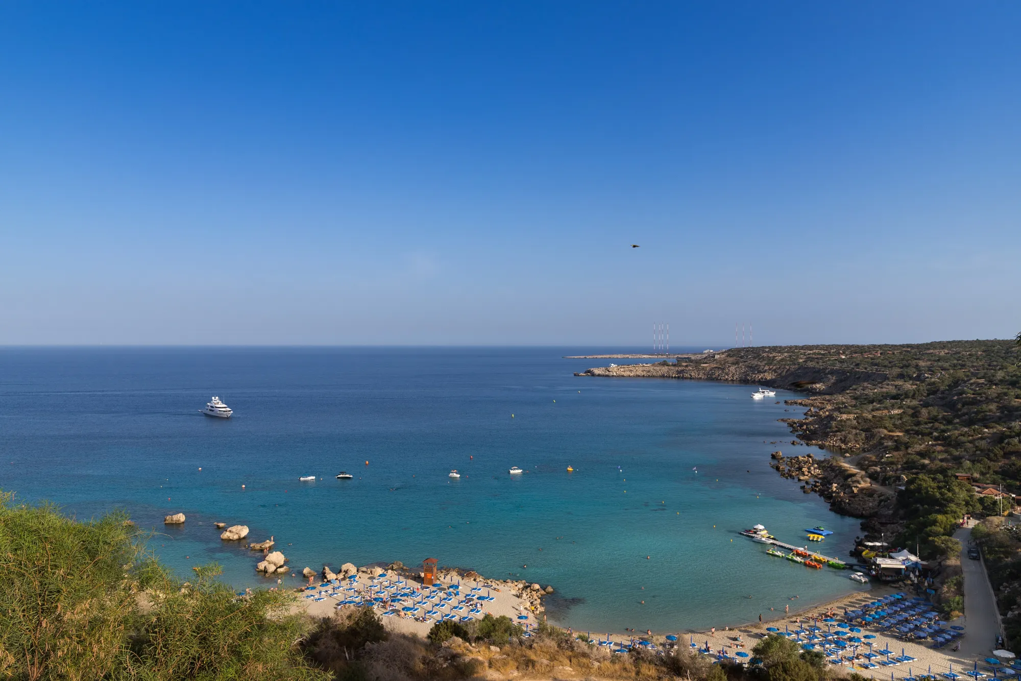 Konnos Bay seen from the road above: a curved sandy cove with rows of blue sunbeds, turquoise shallows, boats anchored offshore and scrub-covered headland on both sides