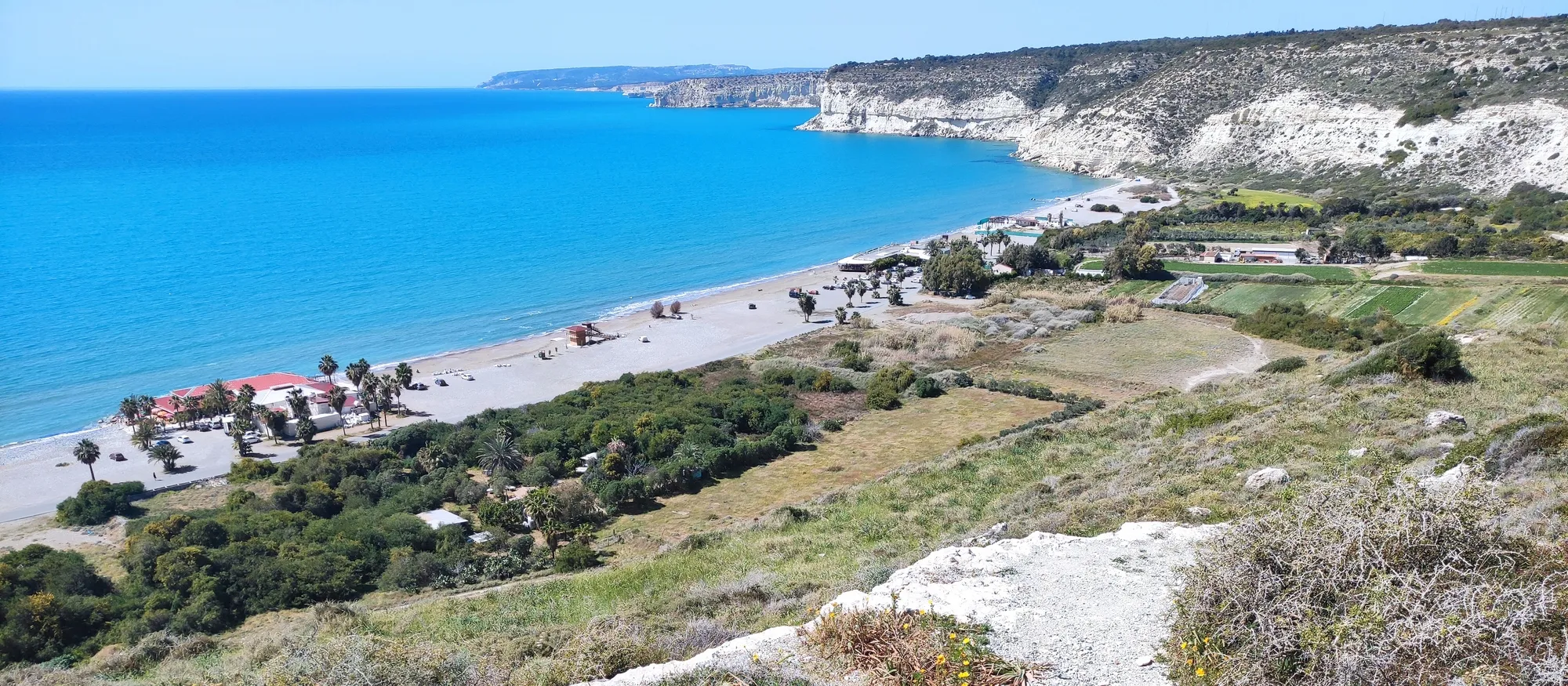 Kourion Beach seen from the clifftop path near the ancient city, with palm-shaded tavernas and sunbed parasols along a pale sand and pebble shore, turquoise water, and white chalk cliffs running west along Episkopi Bay