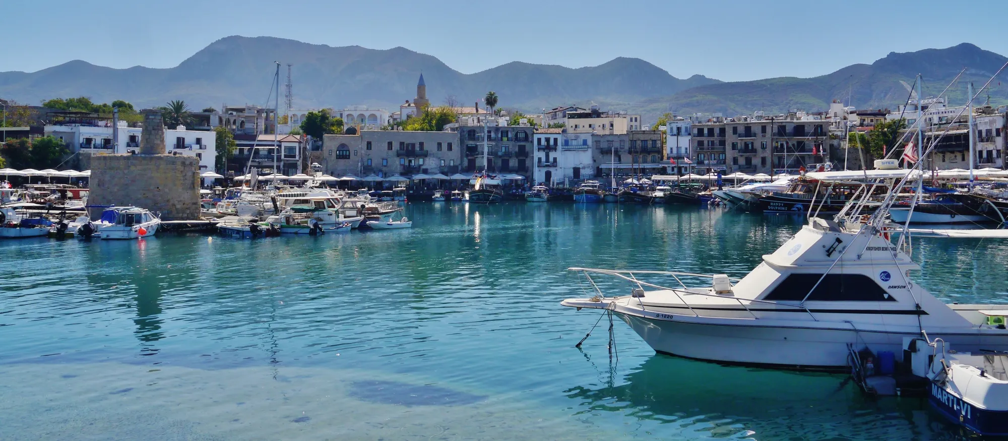 Kyrenia's old horseshoe harbour on a clear day, with turquoise water, moored motor boats and gulets, a row of stone waterfront buildings and the Kyrenia mountain range rising behind the town