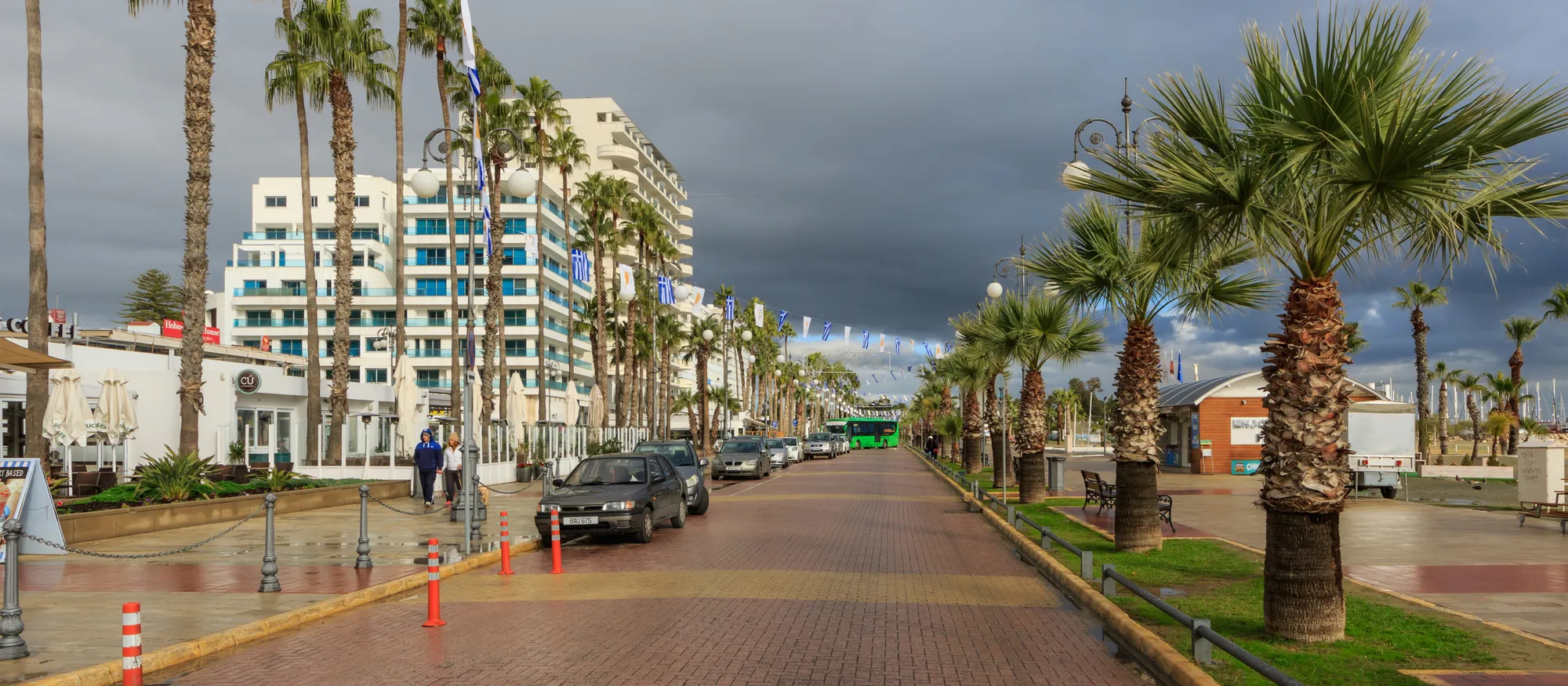 The palm-lined Finikoudes promenade in Larnaca under a dark winter sky, with seafront apartment blocks on the left and a green bus in the distance at the end of the avenue