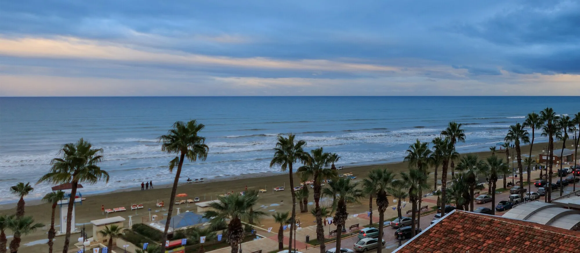 Palm-lined Finikoudes promenade and its sandy town beach in Larnaca, seen from above, with the sea and a cloudy evening sky behind