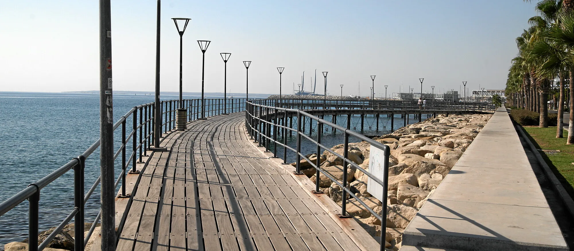 Curving wooden boardwalk along the Limassol seafront, with rock breakwater, palm trees and the masts of Limassol Marina in the distance