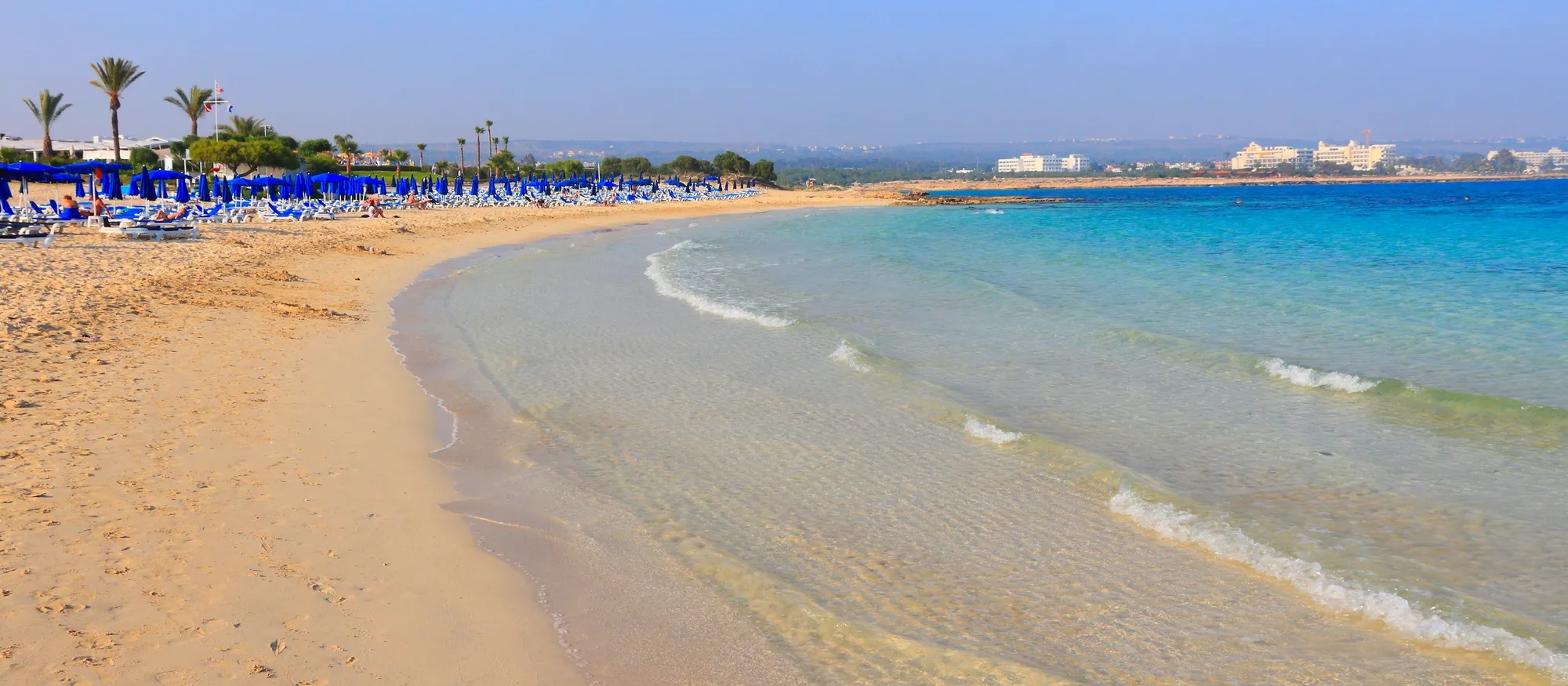 The curved main bay at Makronissos Beach near Ayia Napa, with rows of blue sunbeds and umbrellas on pale golden sand, clear shallow water lapping the shore and low rocky headlands in the distance