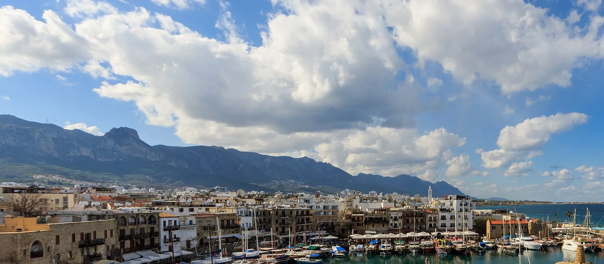 Kyrenia's old harbour seen from the castle bastion, with fishing boats and gulets on turquoise water, stone quayside buildings and the Kyrenia mountains under a blue sky