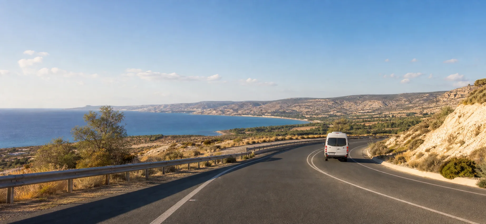 A white airport shuttle travelling on a winding coastal road above the Mediterranean in western Cyprus