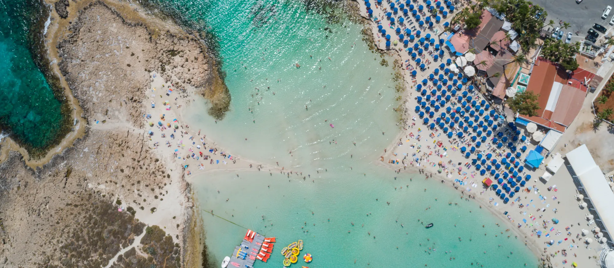 Aerial view of Nissi Beach in Ayia Napa, a pale sandbar joining a low rocky islet to the shore, with turquoise shallows, rows of blue parasols and a pontoon of pedalos