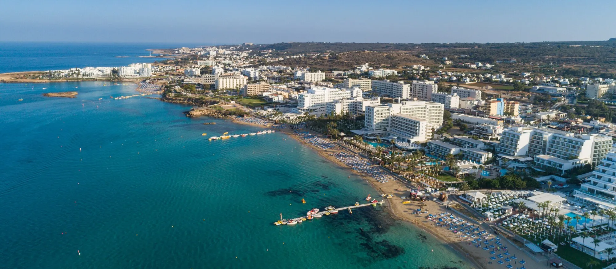 Aerial view of Protaras on the east coast of Cyprus, white resort hotels lining a sandy bay with turquoise water, pontoons and sunbeds in the foreground