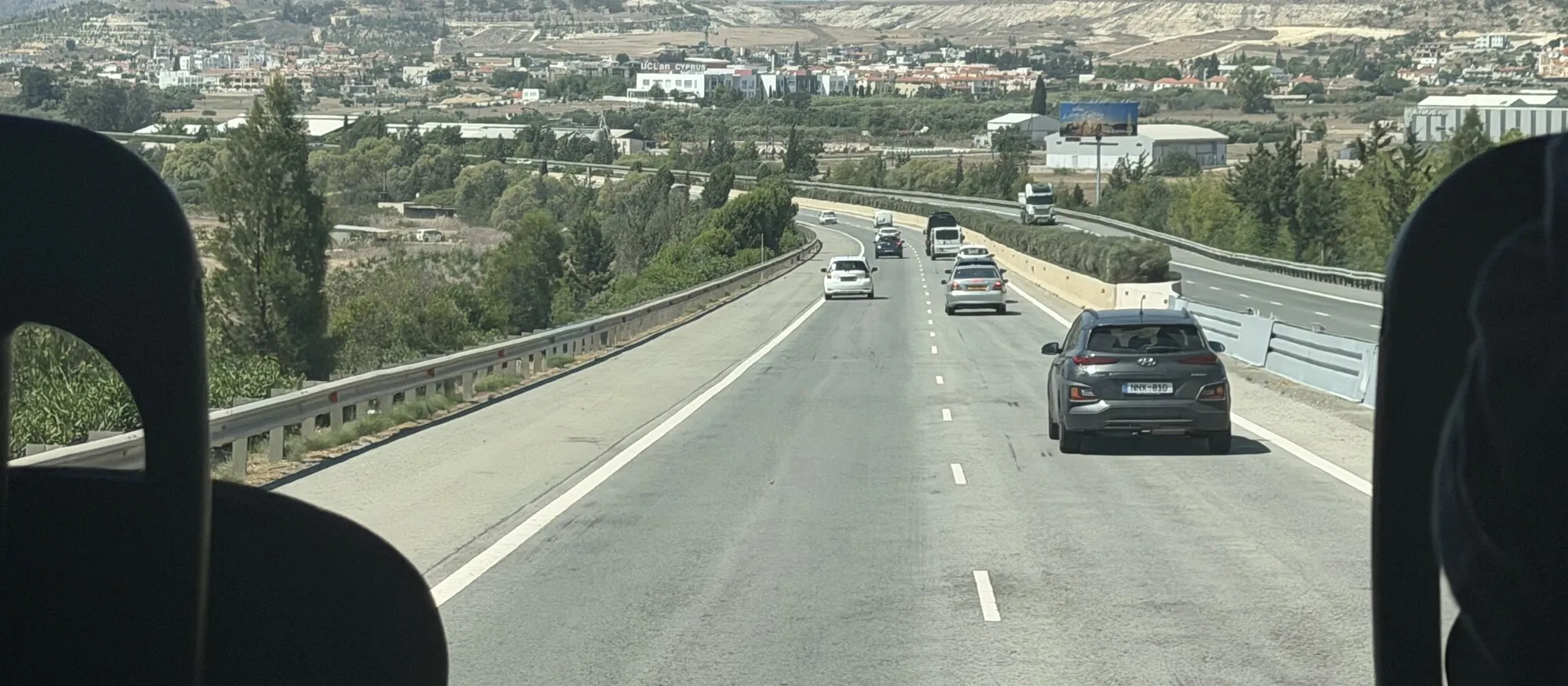View from a coach seat along the A3 motorway near Larnaca, with cars ahead on the dual carriageway, low hills and pale quarried slopes in the distance and the Pyla industrial estate to the left