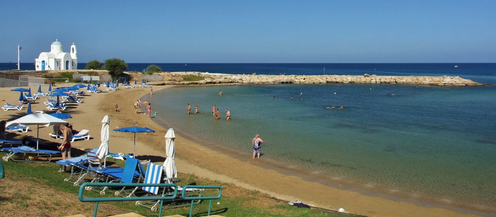 Kalamies bay in Pernera on a clear day, with sunbeds and umbrellas on the sand, a rock breakwater sheltering the bay and the white Agios Nikolaos chapel beside the fishing harbour on the left