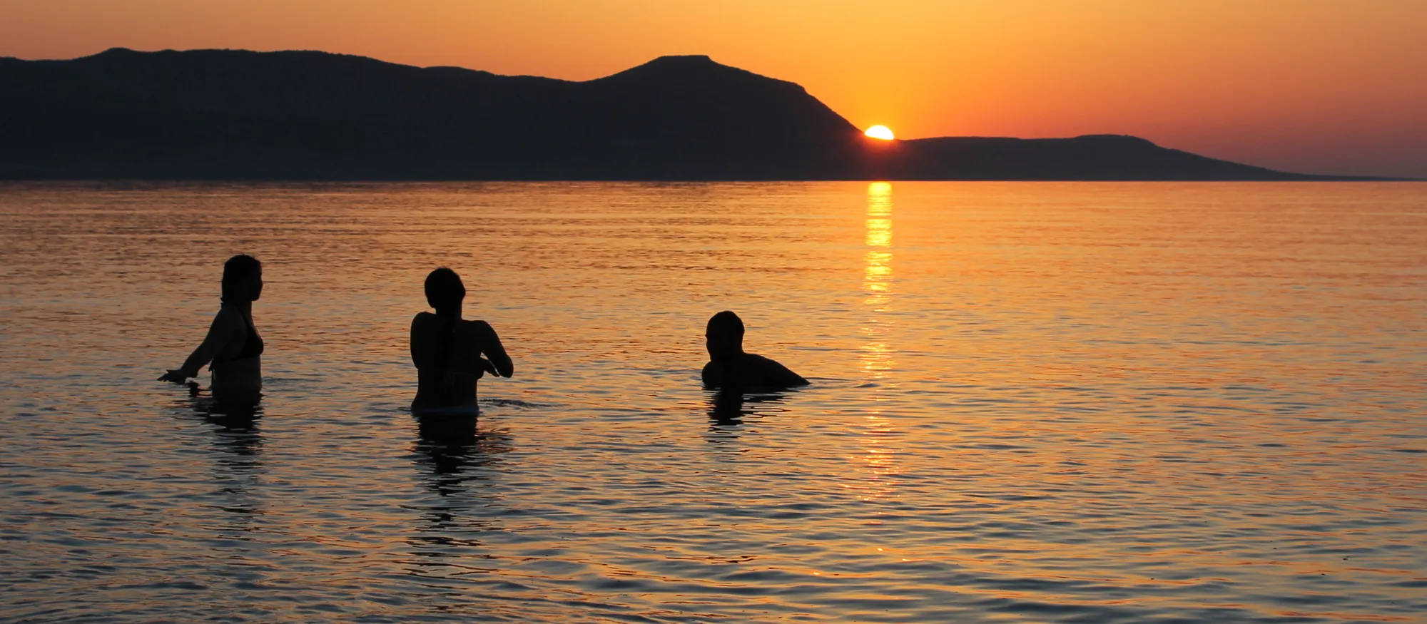 Three swimmers standing in calm shallow water at sunset on the Polis Chrysochous shoreline, silhouetted against the Akamas headland with the sun setting behind it