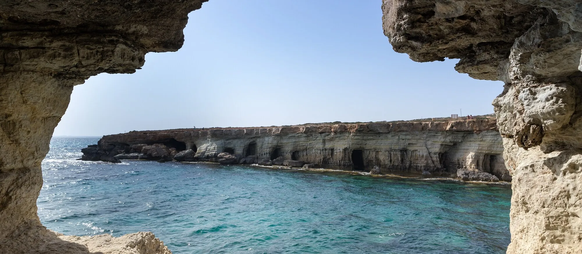 View from inside a limestone sea cave at Cape Greco, Cyprus, looking out across turquoise water to a low cliff line honeycombed with cave openings