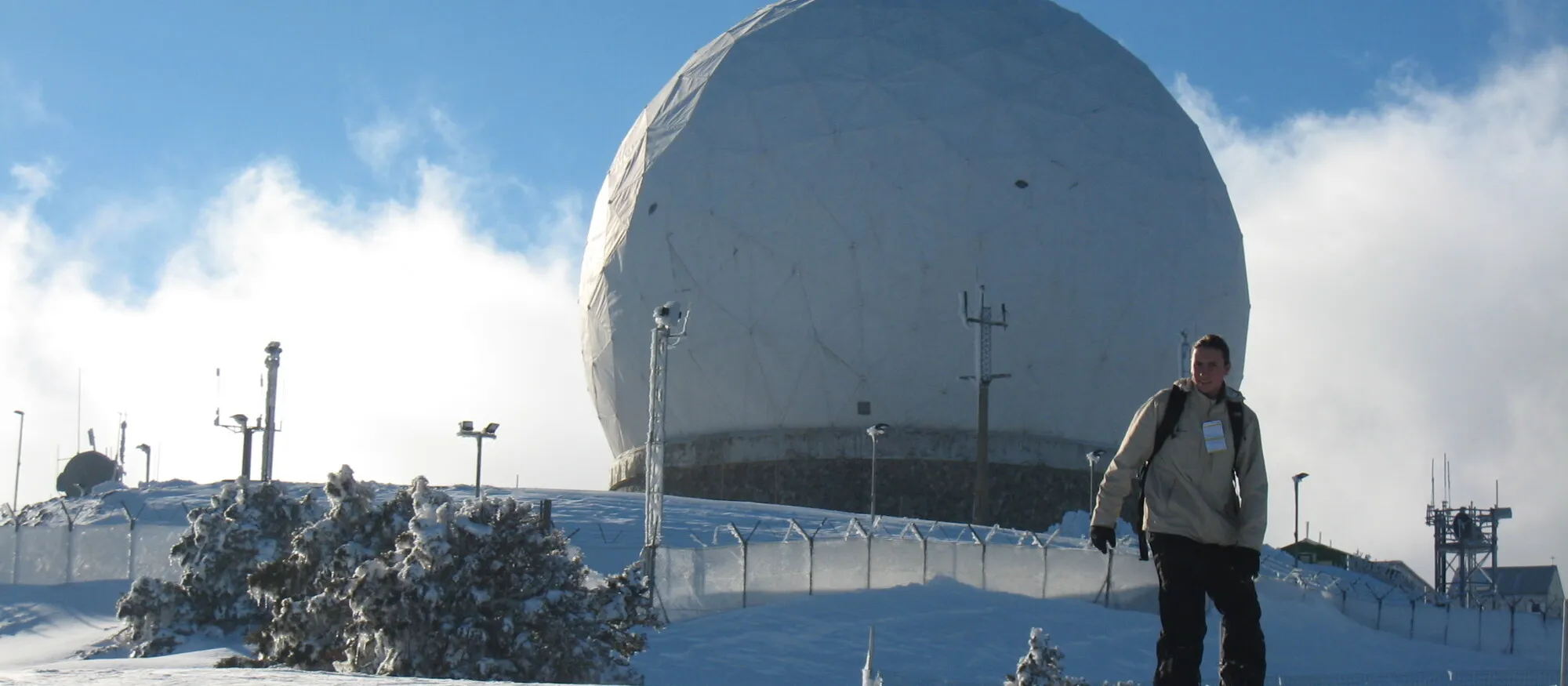A snowboarder standing on packed snow high on Mount Olympus in the Troodos Mountains, with the white radar dome of the summit station behind