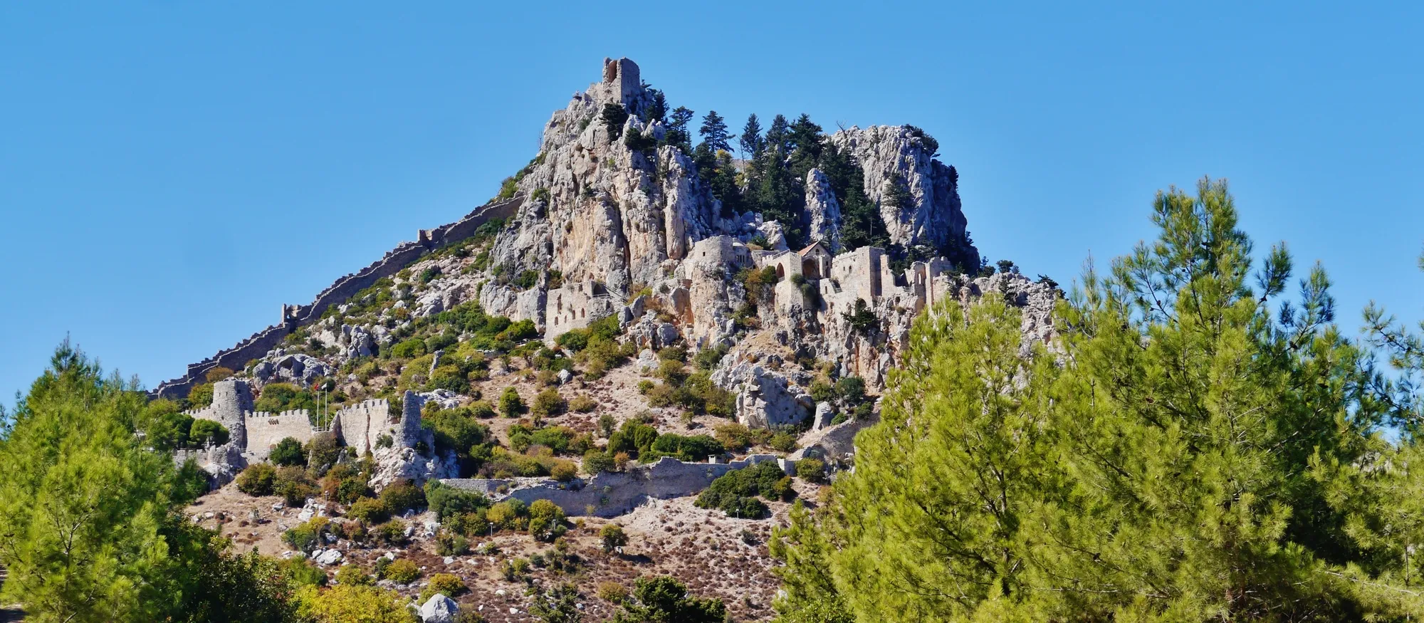 St Hilarion Castle's walls and towers climbing a jagged limestone peak above pine trees, with the lower ward's battlements on the left and the summit tower against a clear blue sky