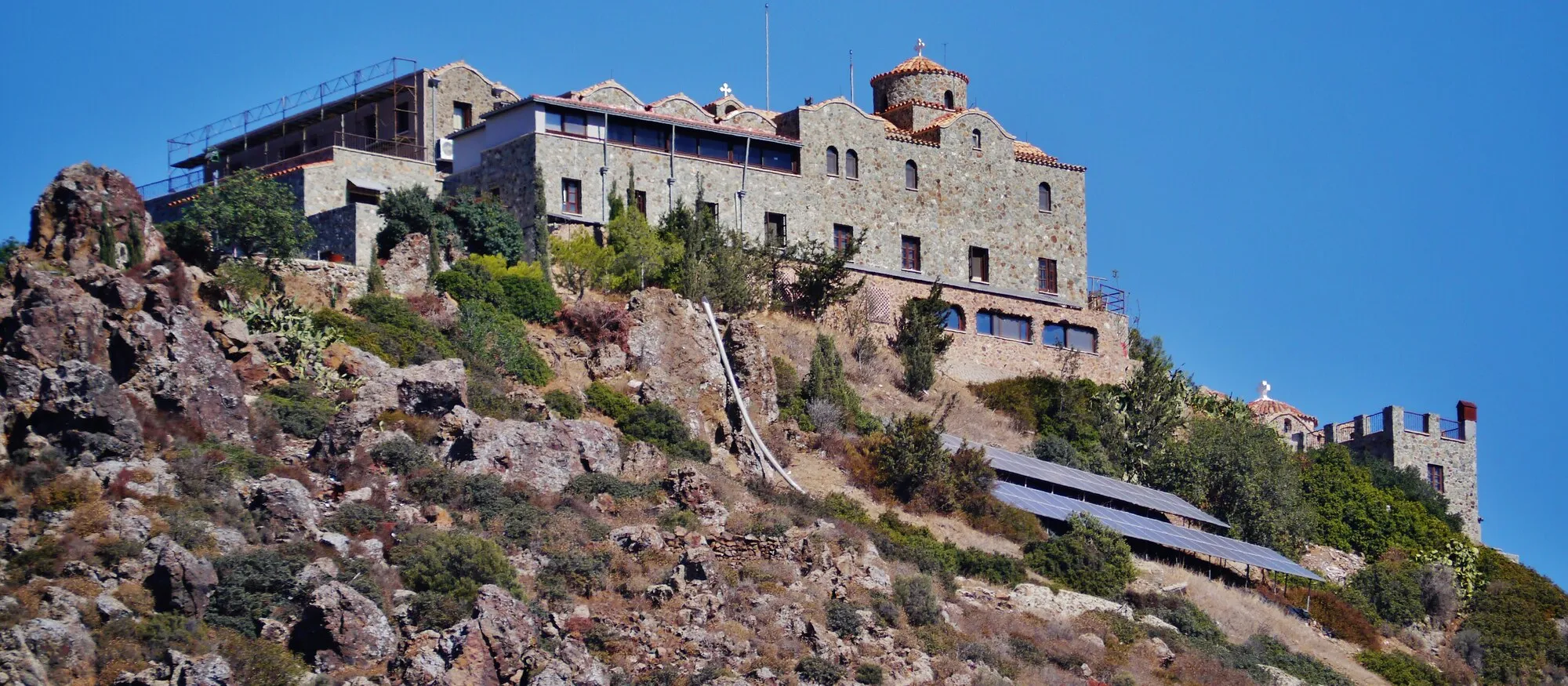 The stone buildings of Stavrovouni Monastery, with a tiled dome and bell cross, standing on top of a rocky, scrub-covered peak under a clear blue sky, with a small crenellated tower lower down on the right