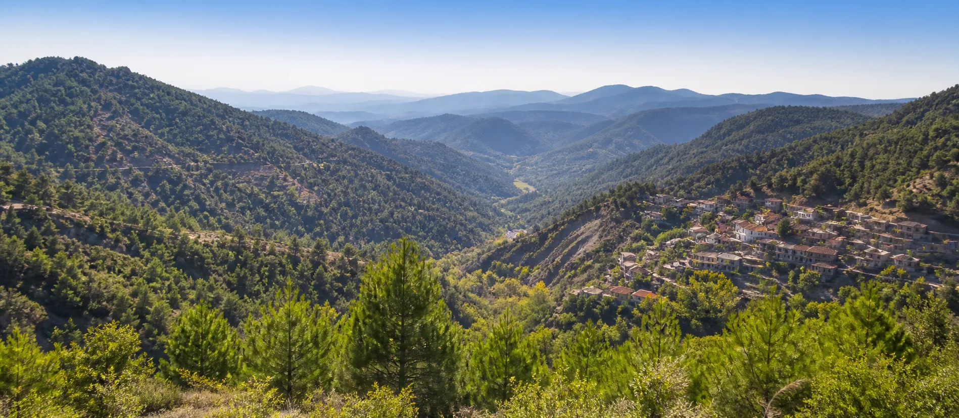 Pine-covered Troodos slopes and a traditional stone village across a Cyprus mountain valley