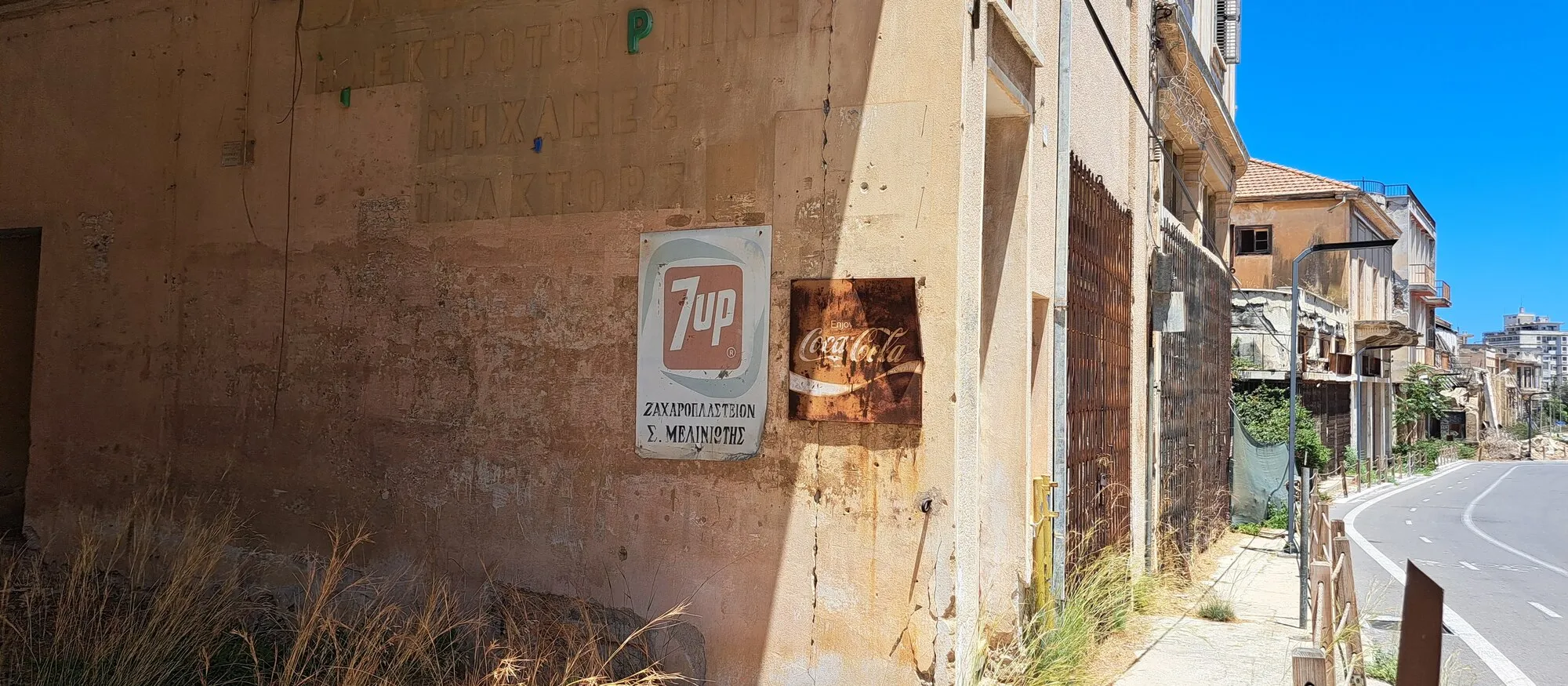 Faded 7up and Coca-Cola signs on the sun-bleached wall of an abandoned Greek Cypriot shop in Varosha, beside a newly paved road with a cycle lane, a wooden barrier and empty buildings under a blue sky